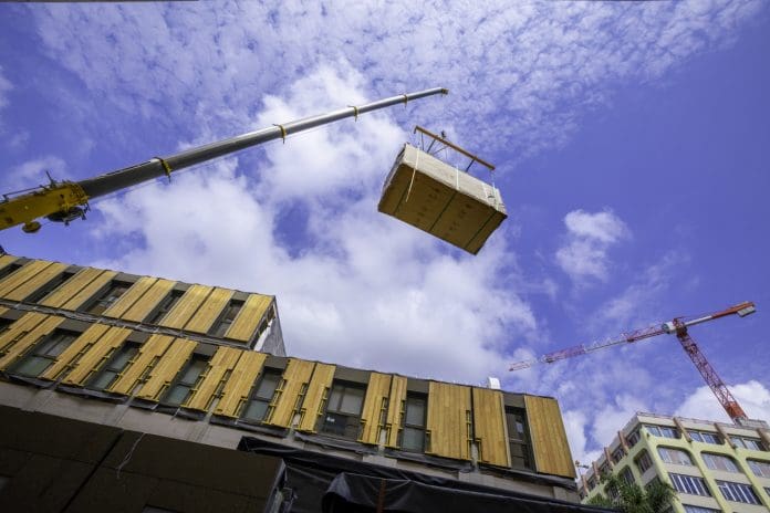 Pre-fabricated modular wood commercial building being assembled with crane A crane with spreader frame lifting a building block of a temporary prefabricated sustainable timber modular office structure in a city environment.