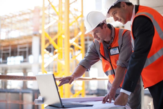 Construction workers using laptop on construction site Two male workers in hi-vis and protective gear using construction software via a laptop on a building site